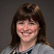 A woman with short brown hair and bangs, wearing a black floral print blouse and gray cardigan smiling in front of a dark gray studio background
